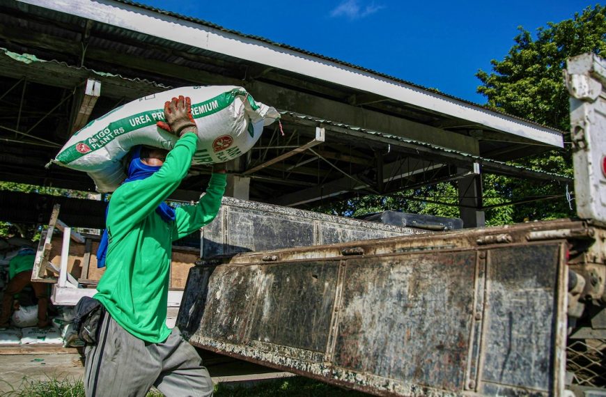 Photo of Man in Green Jumper Carrying a White Bag of urea fertilizer on his Head